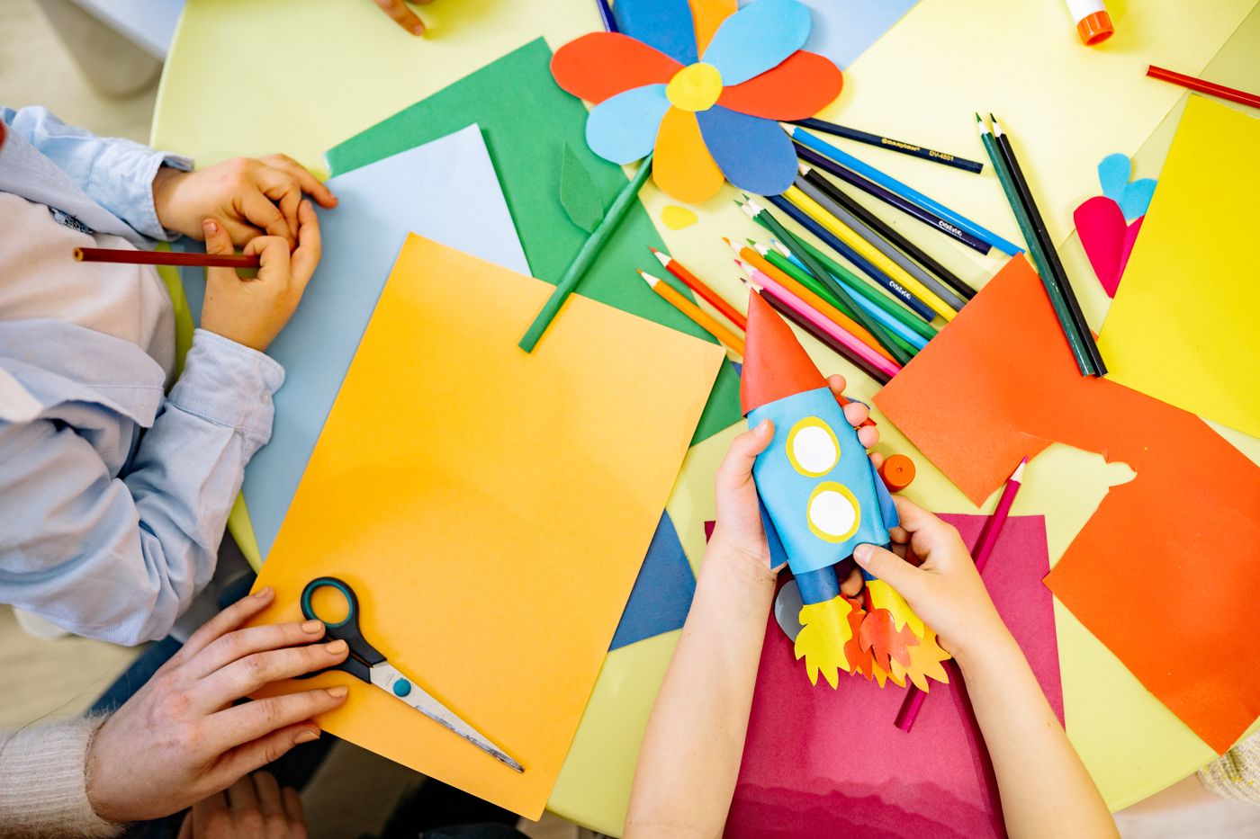 Children's hands making a paper rocket surrounded by coloured paper and pencils