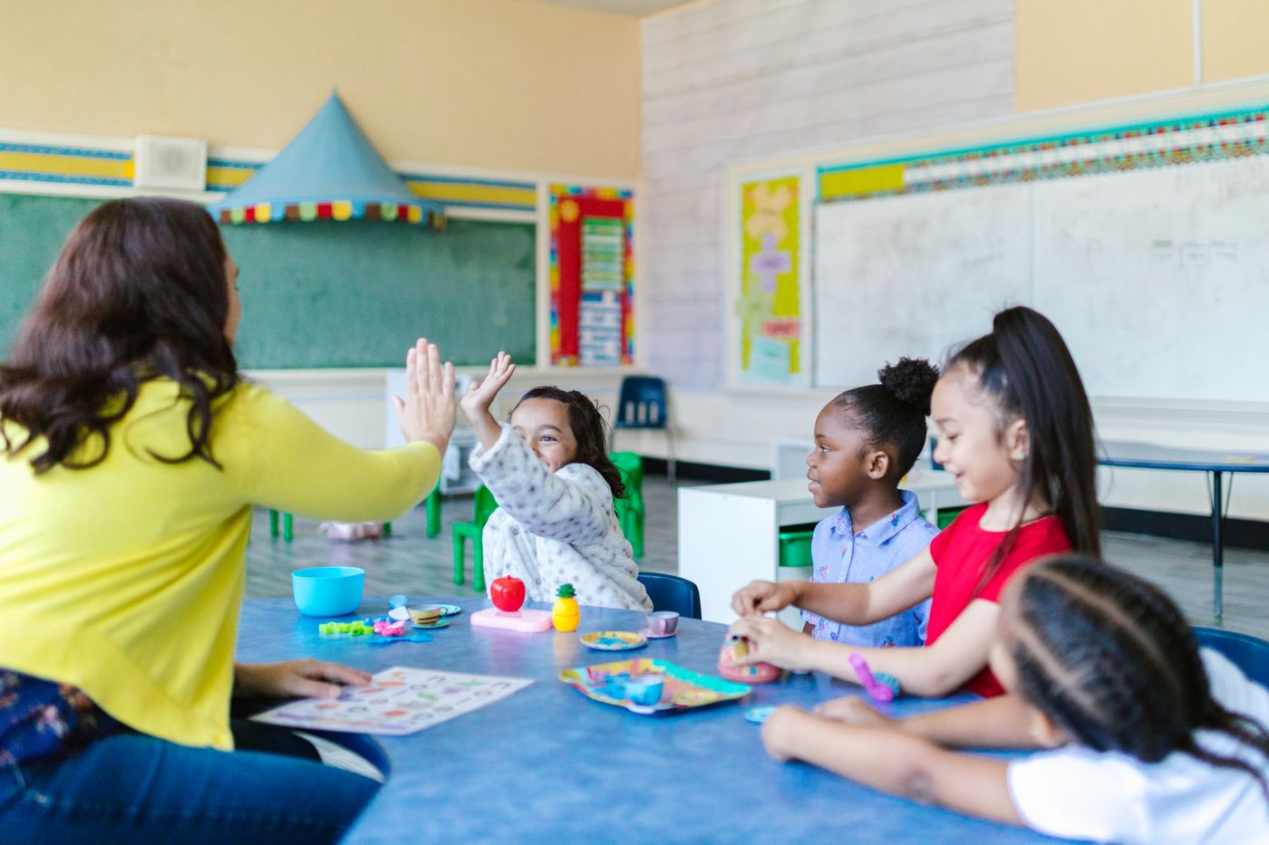 A teacher high-fiving a child across a classroom table while other children play and learn