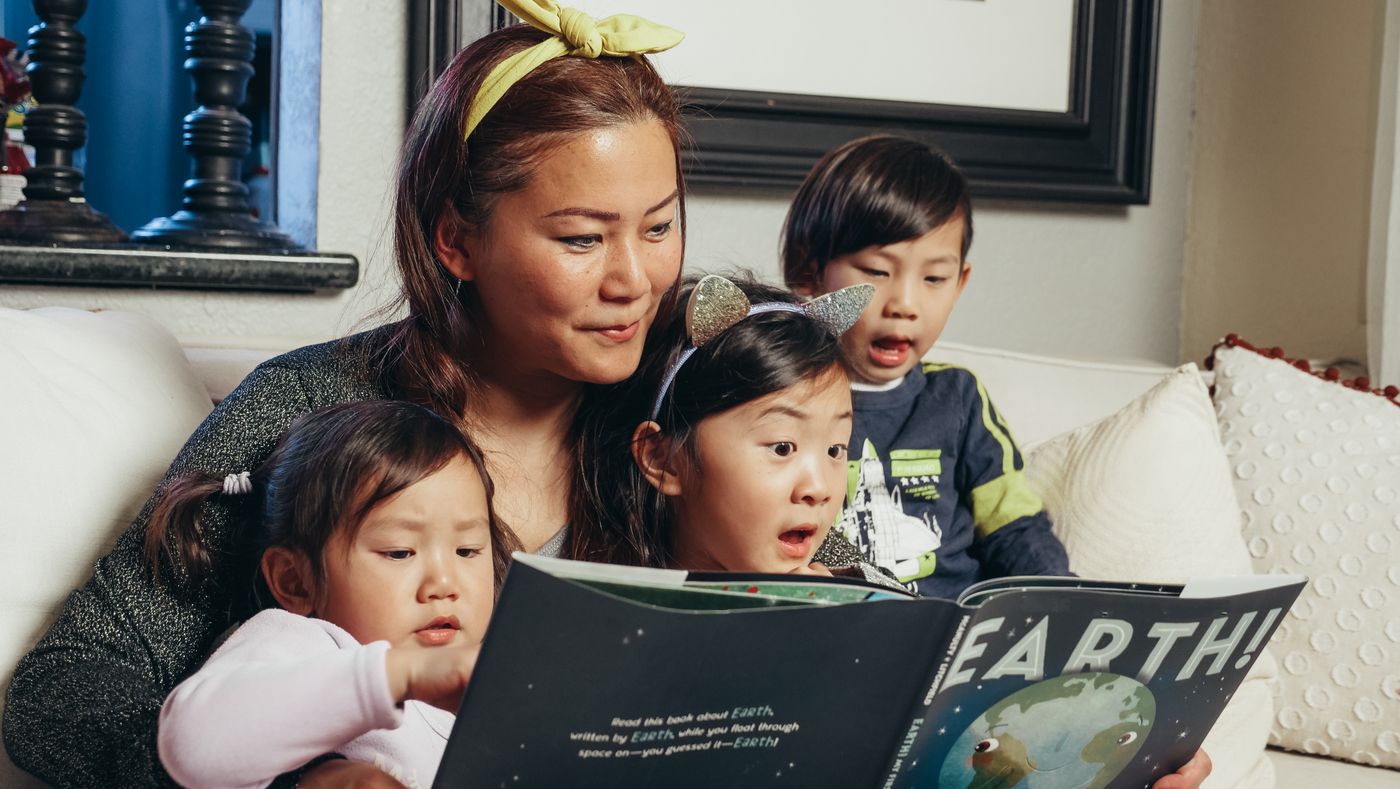 A parent reading a picture book with three young children on a sofa